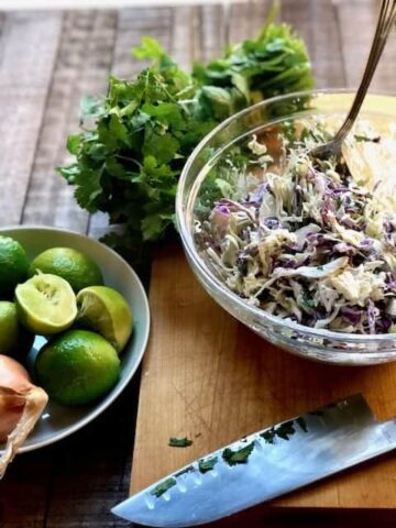 cole slaw in a bowl with a bowl of limes and cilantro next to it