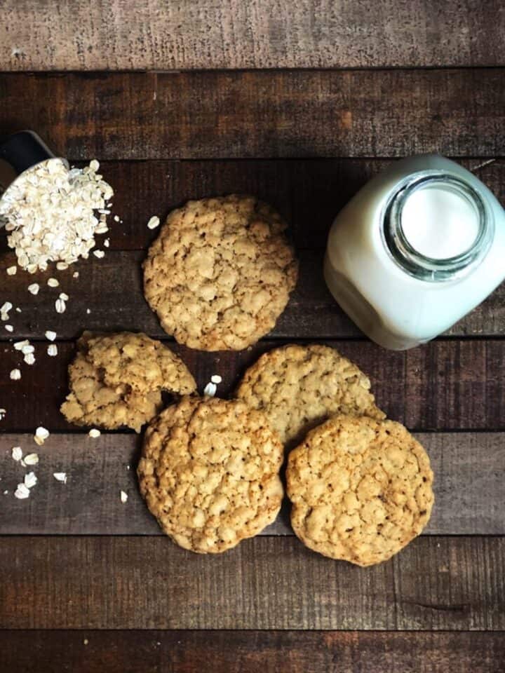 Four oatmeal cookies on a wood board next to old fashioned oats and milk.