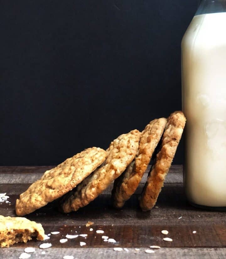 Oatmeal cookies stacked on their side on a wood board and leaning on a pint of milk.