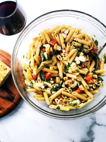 A large bowl of pasta salad on the counter with a glass of wine next to it and bread.