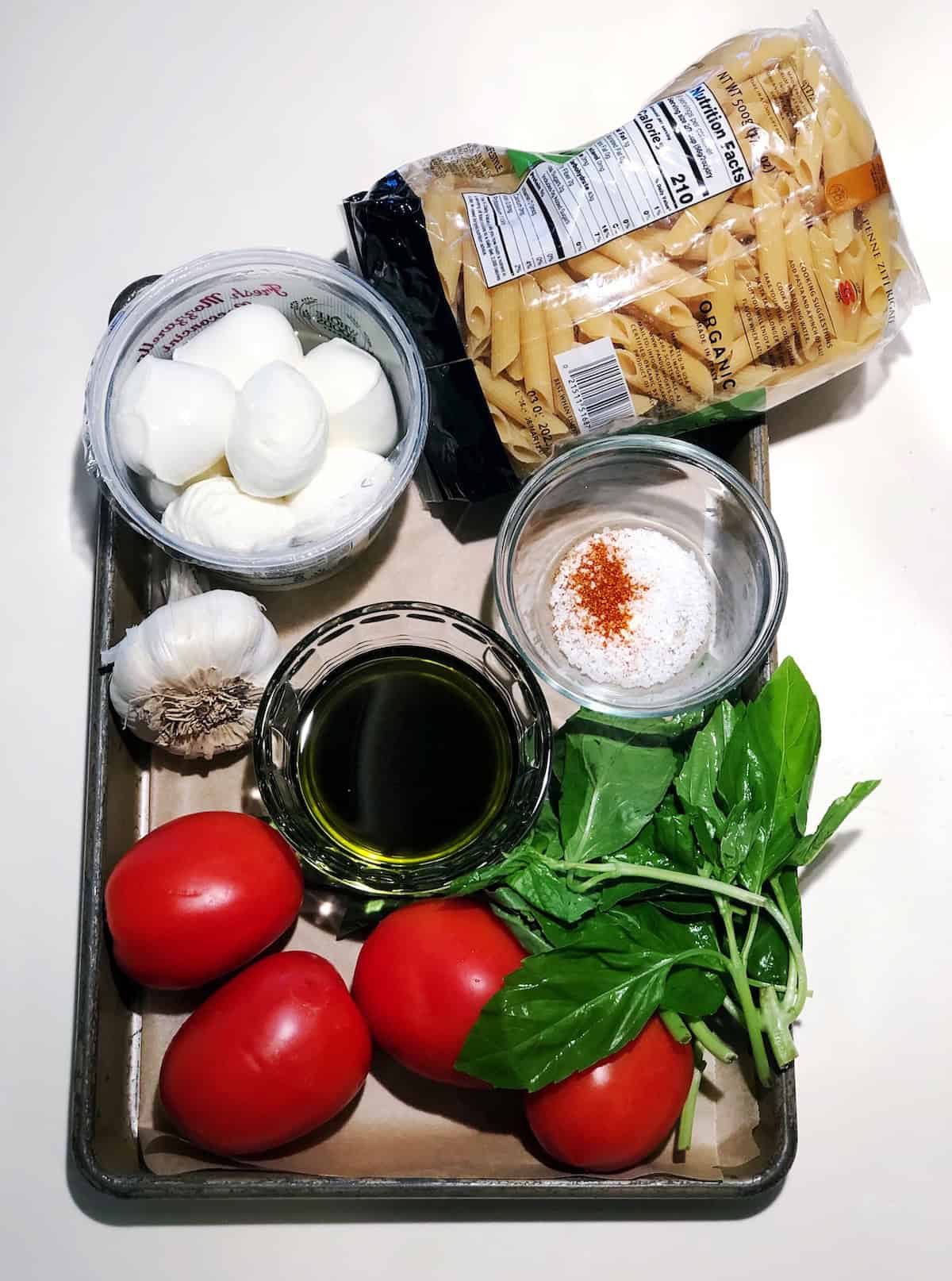Ingredients for the pasta salad on a tray including tomatoes, garlic, pasta, basil, olive oil and balsamic vinegar, spices in a small bowl and fresh mozzarella.
