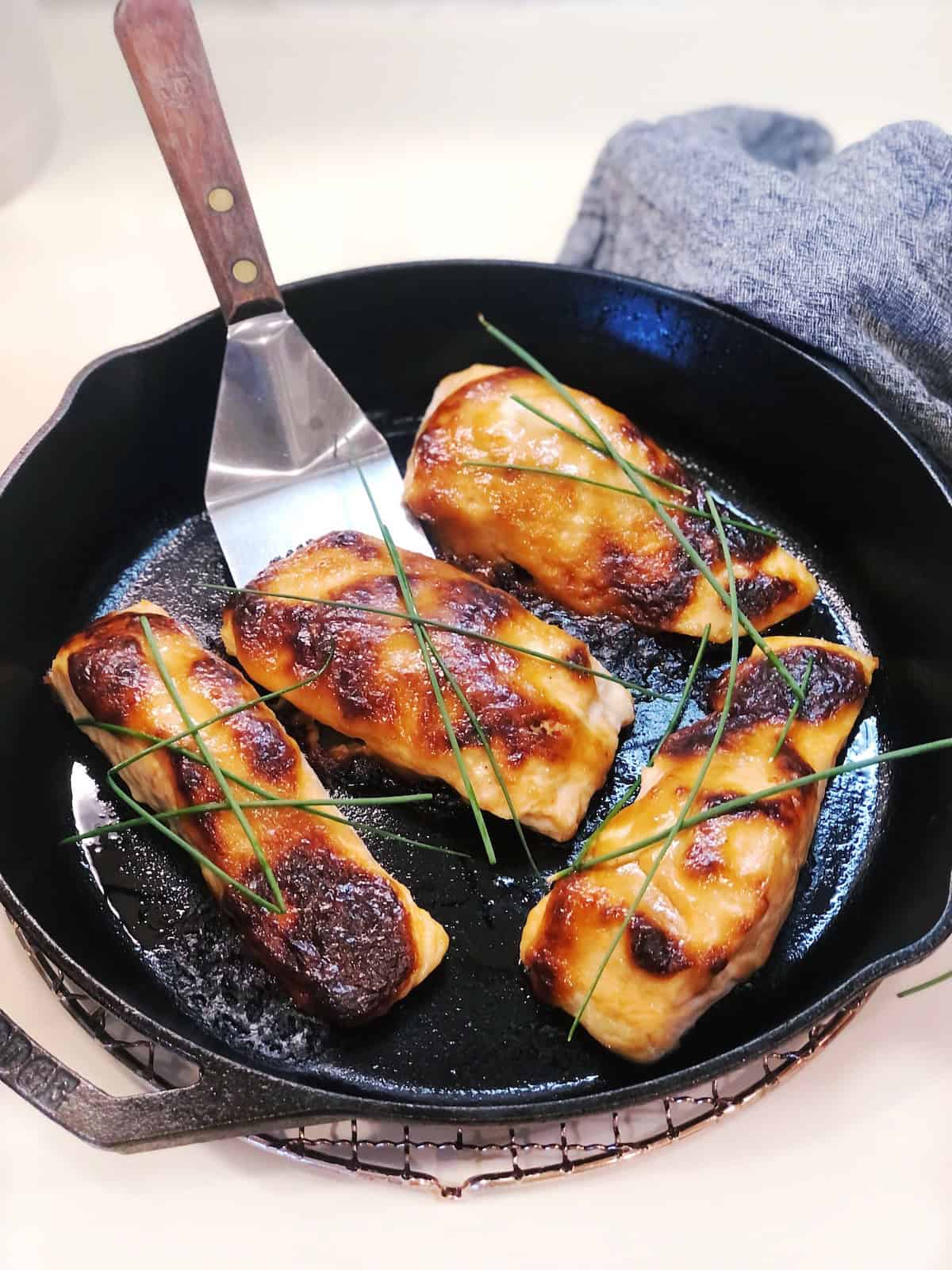 Four baked miso salmon filets in a cast iron pan with a spatula in the pan and chives on top.