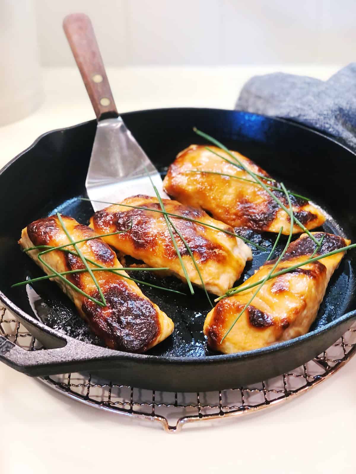 A zoomed in view of four baked miso salmon filets in a cast iron pan with a spatula in the pan and chives on top sitting on the counter.
