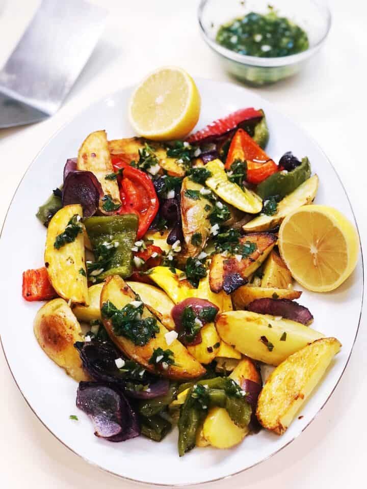 The roasted vegetables with green chimichurri sauce on top of them on a large oval plate with extra chimichurri sauce in a bowl in the background.