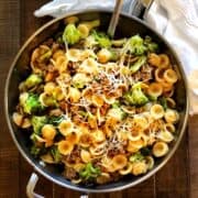 Orecchiette with sausage and broccoli in a large saute pan on a wood table with a cloth napkin next to it.