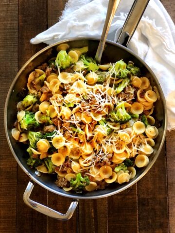 Orecchiette with sausage and broccoli in a large saute pan on a wood table with a cloth napkin next to it.